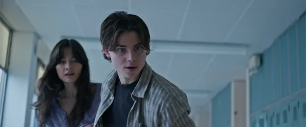 A still from the film This Is Not A Test. A white teenage boy and girl look downwards with disturbed expressions as they make their way along a school corridor.