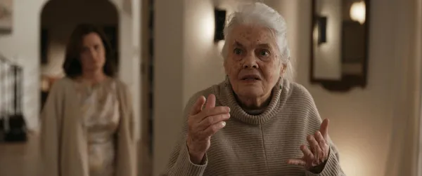 A still from the film The Uninvited. An older white woman with an intense yet bewildered expression holds up her hands in front of her as she wanders through spacious rooms. A younger white woman is out of focus behind her.