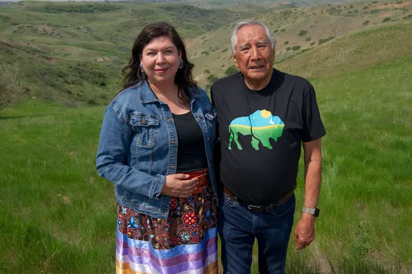A Cree woman and an older Cree man with rolling green hills stretching out into the distance behind them.