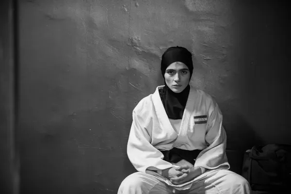 A black and white still from the film Tatami. An Iranian woman in a headscarf and judo robe sits croo-legged in the corner of a small room.
