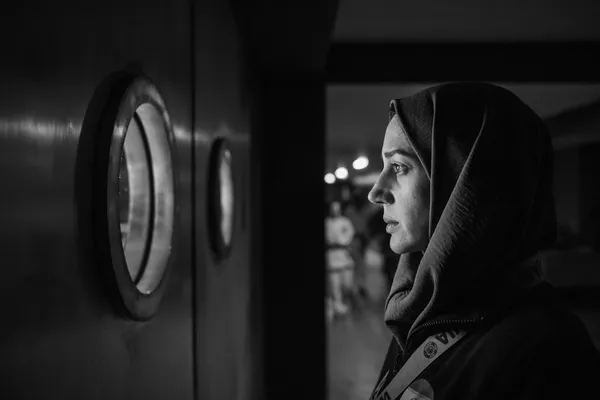 A black and white still from the film Tatami. An Iranian woman, wearing a headscarf, stands in a corridor looking out through a round window.