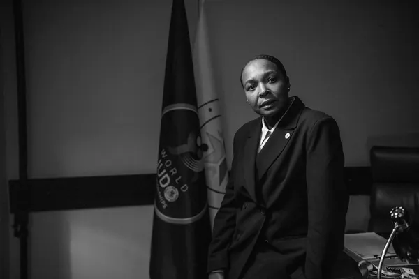 A black and white still from the film Tatami. A short-haired Black woman in a black suit leans against a desk. There is a flag behind her bearing the words 'world judo'.