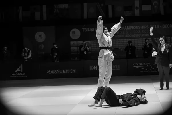 A black and white still from the film Tatami. An Iranian woman in a headscarf and white judo robes stands, triumphant, over a prone woman in dark judo robes, in an arena.
