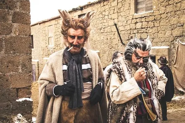 A still from the film The Time It Takes. Two middle-aged white men in elaborate costumes, with wild hair, eyebrows and moustaches, stand on a corner among low stone buildings, smoking cigarettes.