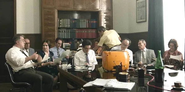 A still from the film The Great Ambition. Ten white people sit around tables looking at pieces of paper.