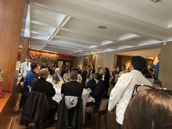A group of white man sit around a table while other white people crowd in around the edges of the large room.