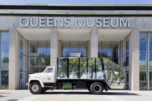 A mirrored truck is parked outside the glass doors of a building marked Queens Museum.