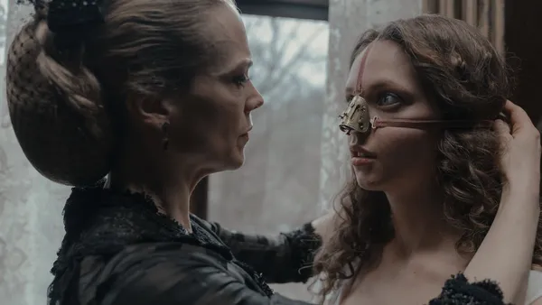 A still from the film The Ugly Stepsister. An middle aged woman in old fashioned clothing straps a device with a metal nose-brace over a young woman's face.