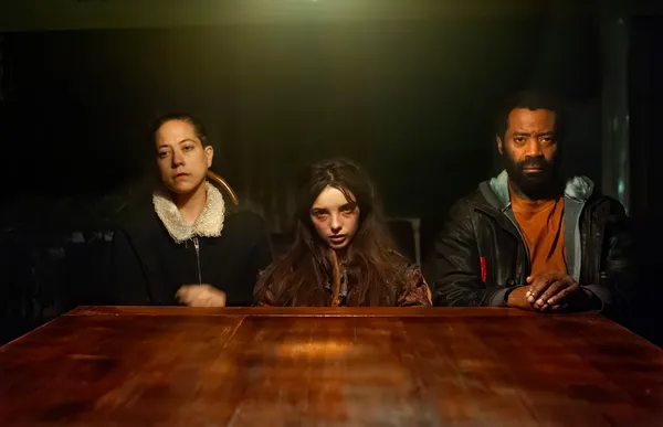 A still from the film The Wolf, The Fox And The Leopard. Across a wooden table in a dark room, a white woman and a Black man flank a white teenage girl with long, tangled hair and a bruised face.