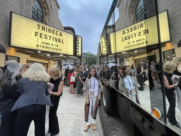 A light-skinned woman with a lanyard around her neck stands by the side of a shiny truck, which is parked opposite a tall sandstone building with a sign saying 'Tribeca Festival'.