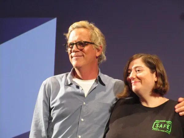 A bespectacled middle-aged white man with white hair, wearing a pale grey shirt, stands with his arms around a shorter, brown-haired white woman wearing black.