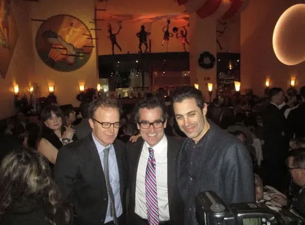 Three light-skinned men, wearing suits, stand together in a crowded bar.