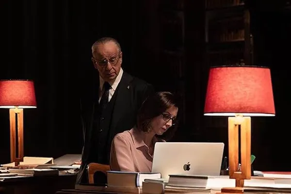 A still from the film La Grazia. A older white man looks over the shoulder of a white woman who is working on a laptop in a dark room.