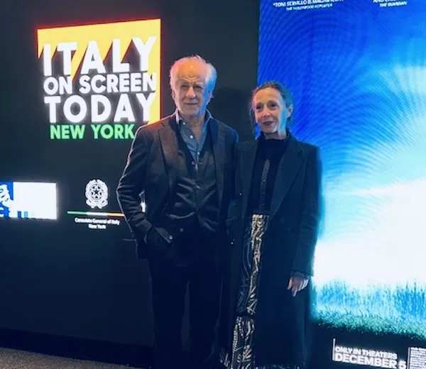 An older white man and a middle-aged white woman stand beside a sign which reads 'Italy on screen today, New York'