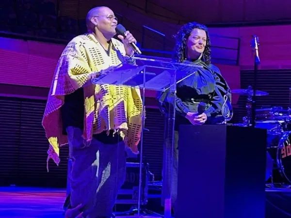 Two Black women, one with a shaven head and yellow poncho, the other with long hair and dark clothing, stand on a stage illuminated by purple light.