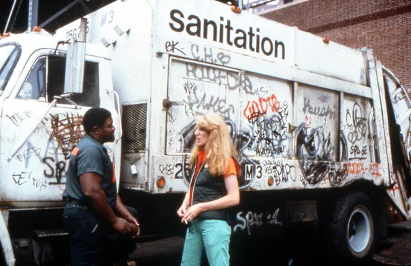 A Black man and a blond white woman, both wearing blue collar work clothes, are talking beside a white van marked 'sanitation', which is covered in graffiti.