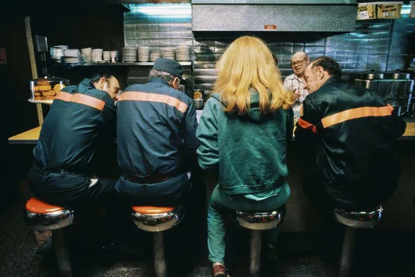 Three men in sanitation workers' uniforms and a blond woman, seen from behind, sit at a café counter.