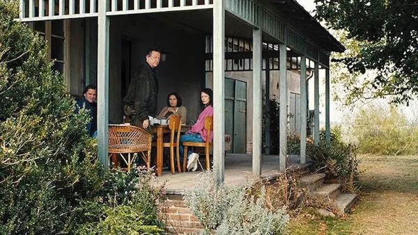 A still from the film Mirrors No. 3. Four white people gather around a table on a veranda in a spacious garden.