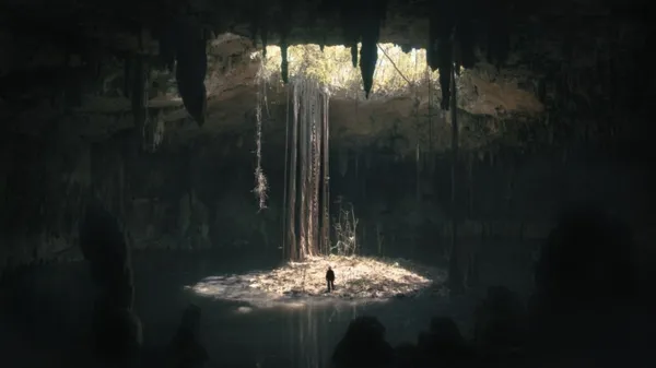 A still from the film Underland. A human figure stands on a small island in the centre of a flooded underground cavern full of stalactites and stalagmites. Light travels down from an opening directly above, through which foliage can be seen.