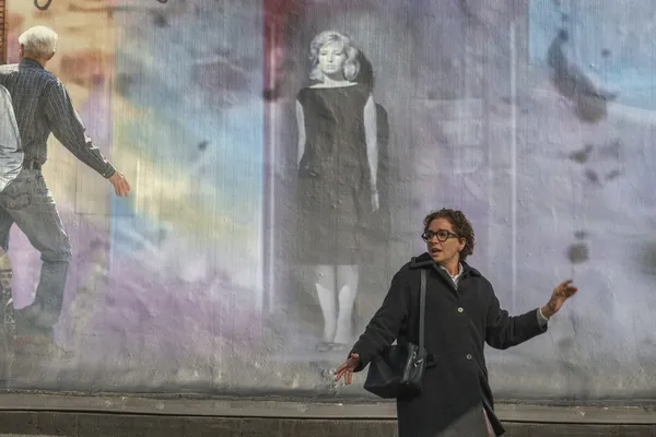 A still from the film La Gioia. A bespectacled middle-aged white woman with dark hair looks back, gesticulating expressively, as she passes a mural which features a black and white image of a slim blonde in a little blacl dress.