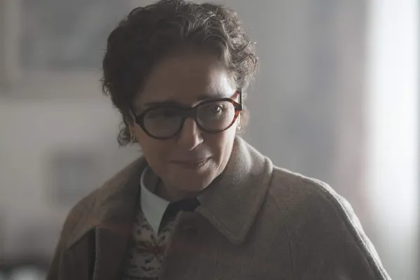 A still from the film La Gioia. A bespectacled middle-aged white woman with dark hair and a grey coat.