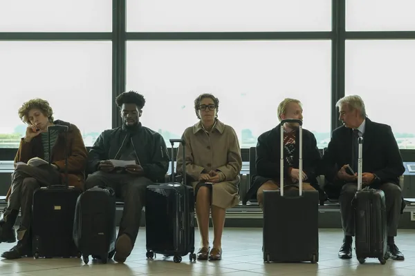 A still from the film La Gioia. Five people with luggage sit in a row in front of big windows at an airport. In the middle is an unhappy-looking bespectacled middle-aged white woman. The sky outside is damply grey.