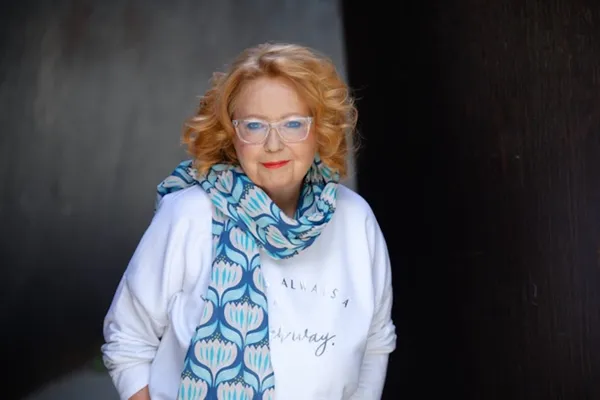 A bespectacled white woman with golden hair, a white coat and a blue and white patterned scarf.
