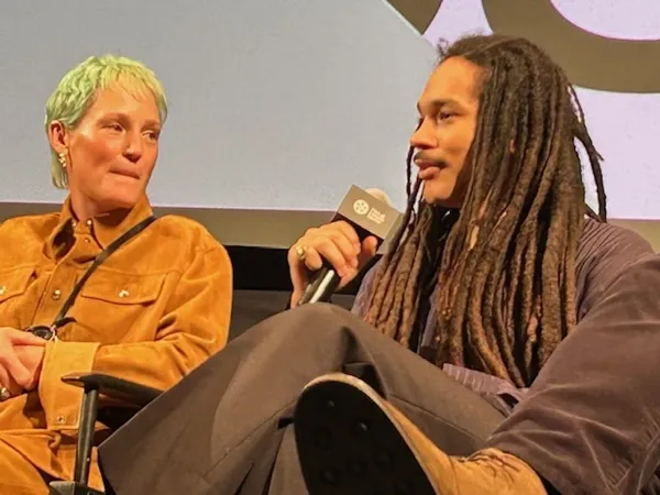 Vicky Krieps and Luka Sabbat. A white woman with light green hair and a mustard velvet suit sits beside a Black man with dark clothes, long dreadlocks and a microphone.