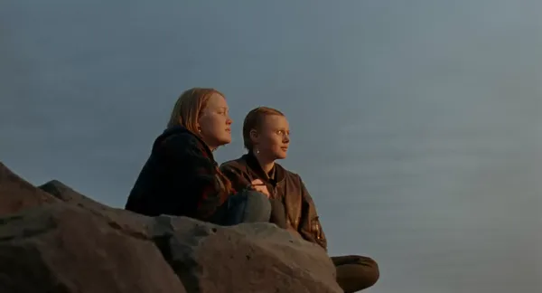 A still from the film When The Light Breaks. Two young white women sit together on a rocky clifftop.