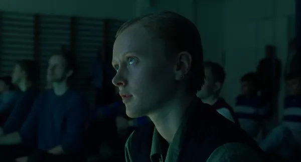 A still from the film When The Light Breaks. A depressed-looking short-haired young white woman sits on the floor of a dimly-lit gym with a group of her peers.