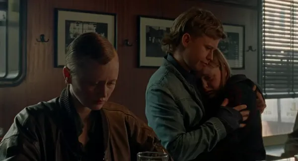 A still from the film When The Light Breaks. A short-haired young white woman sits at a bar looking glum as a young white man in a denim jacket hugs the tearful young white woman beside her.