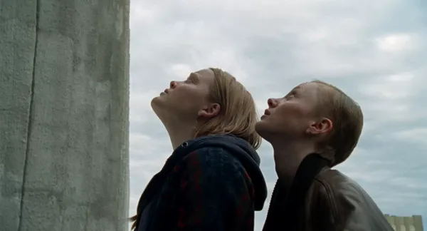 A still from the film When The Light Breaks. Two young white women stand beside a whitewashed stone wall, looking up.