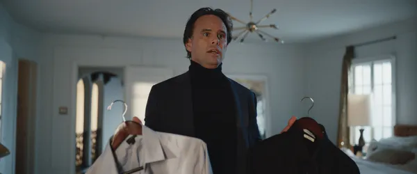A still from the film The Uninvited. In a white suburban bedroom, a white man with a querulous and slightly frantic expression holds up a white suit in his right hand and a black suit in his left hand.