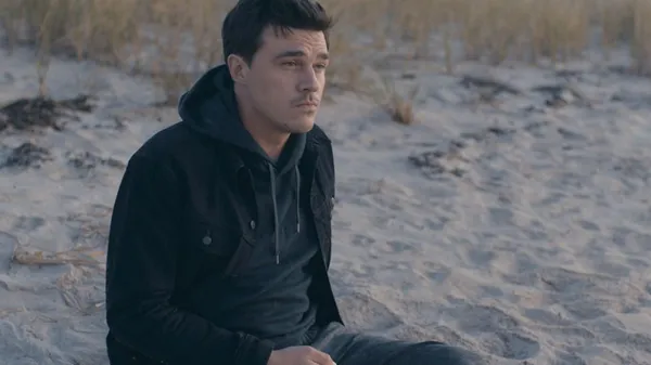 A still from the film Westhampton. A young white man with dark hair and a dark jacket sits alone on a beach, with grass pushing through the sand behind him.