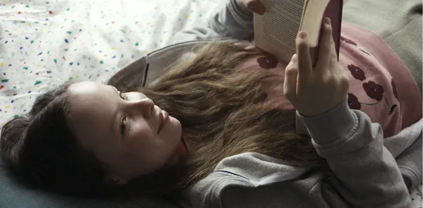 A still from the film What Marielle Knows. A smiling white girl aged about 12, with long light brown hair, lies on a bed reading a book.
