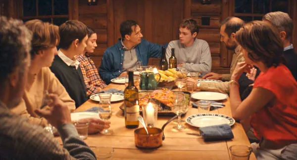 A still from the film Who By Fire. A group of white people sit around a dinner table in the evening.