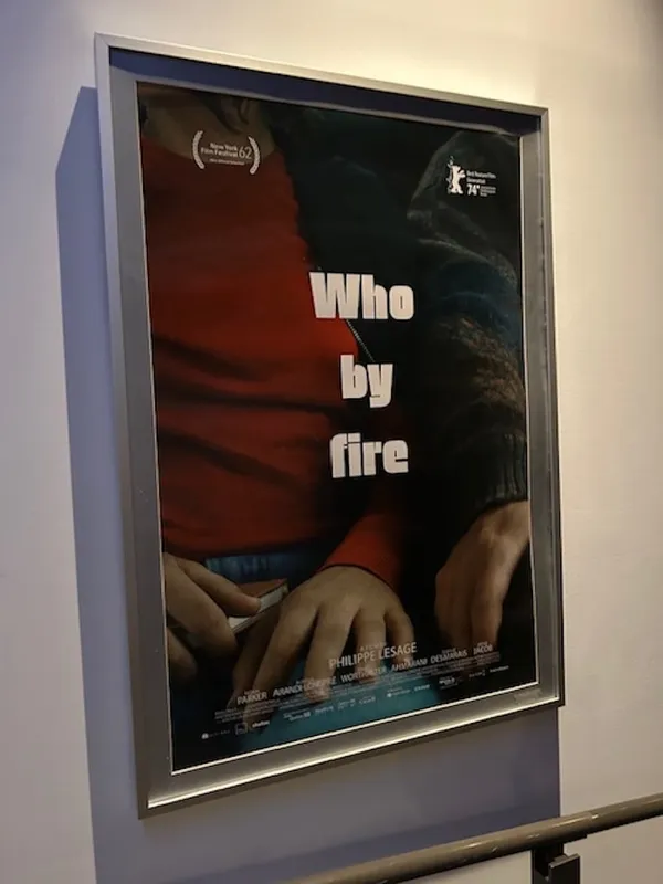 A framed poster for the film Who By Fire, hanging on a white wall.