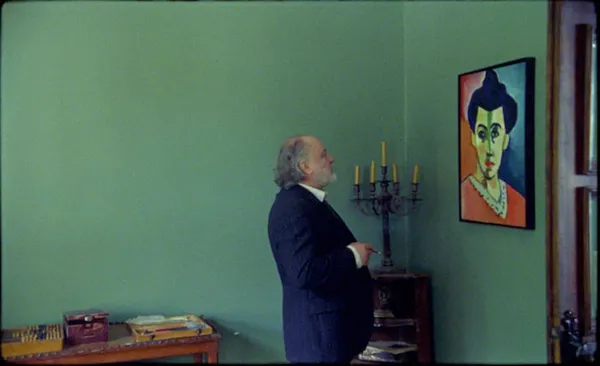 A still from the film The Assistant. In a room with mint green walls, an older white man in a dark suit, holding a cigarette, regards a painting of a pale figure with a line of shadow down the middle of the face.