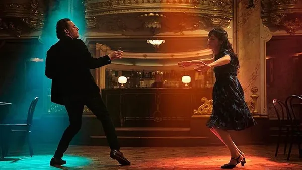 A still from the film Dead Language. A white man in a black suit and a white woman in a black and blue dress dance on a wooden floor in front of a baroque-styled bar.