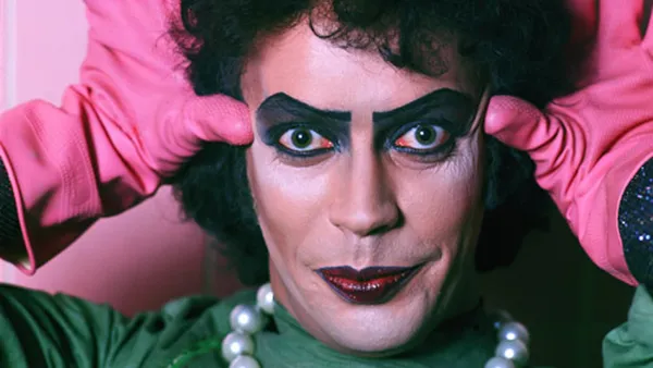 A still from the film The Rocky Horror Picture Show. A close-up on the face of a sweet transvestite wearing a green dress and pink rubber gloves.