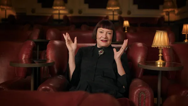 A still from the film Strange Journey: The Rocky Horror Story. An older white woman dressed in black, with a sharply cut bob of auburn hair, sits in a cinema, hands raised expressively.