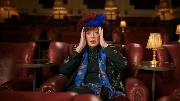 A still from the film Strange Journey: The Rocky Horror Story. A fabulously dressed older white woman whose big red curls are bound up with a blue silk scarf sits in a cinema, one palm against either side of her face.