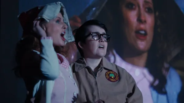 A still from the film Strange Journey: The Rocky Horror Story. A young white woman holds a newspaper over her head beside a young white person wearing glass and a cub scout uniform, in front of a cinema screen.