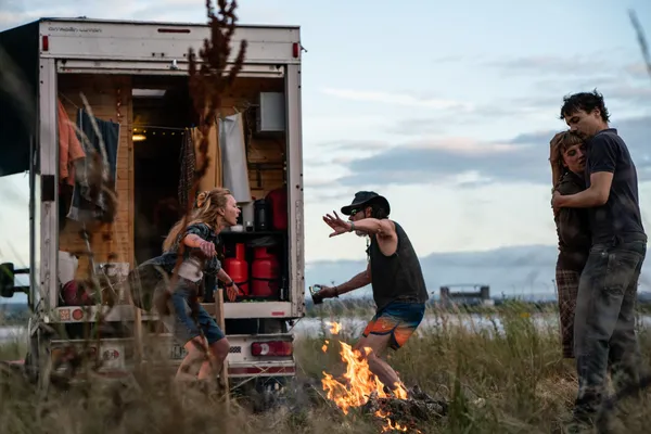 A still from the film Urchin. Two white people dance around a campfire, by daylight, beside a van whose back doors are open, while two others hug.