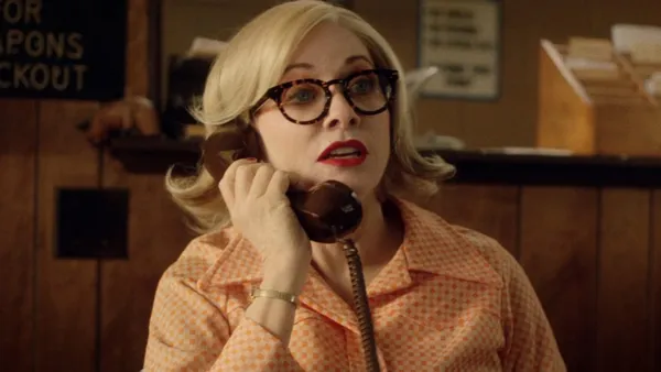 A still from the film The Last Stop In Yuma County. In a wood-panelled office, a white woman with blond bobbed hair, red lipstick and black-framed glasses answers a rotary-dial telephone.