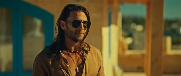 A still from the film Compulsion. A long-haired white man with stubble and sunglasses stands in the portico of a golden sandstone villa. Green hills are visible in the distance, under a blue sky.