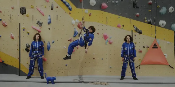 A still from the film Doppelgängers. Three woman wearing royal blue jumpsuits scale a climbing wall.