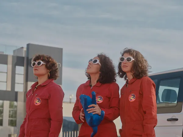 A still from the film Doppelgängers. Three pale, femining figures in red jumpsuits and whire-framed glasses walk past modern buildings. One of them is looking skywards.