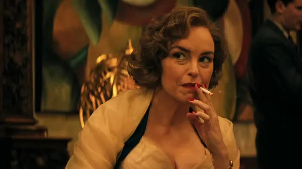 A still from the film Hedda. A middle-aged white woman in a white silk bodice, with an expression of curiosity and amusement, smoking a cigarette.