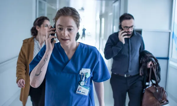 A still from the film Late Shift. A white nurse walks along a white corridor, speaking on her phone. There are other people behind her, one of whom is using his phone.